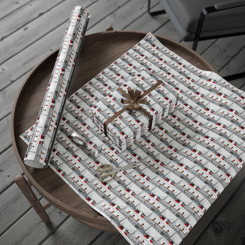 K&ouml;nnte beinhalten: Eine Rolle wei&szlig;es Geschenkpapier und ein Geschenk mit braunem Band, beide mit einem sich wiederholenden Muster aus grauen und roten Kreuzfahrtschiffen. Goldene Schere und Klebeband liegen auf dem Holztisch.