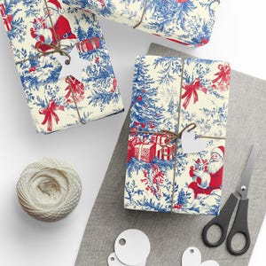 May include: Wrapping paper featuring a repeating toile pattern in red and blue on a cream background, depicting Santa Claus, Christmas trees, and gift boxes. The paper is tied with brown twine and includes white star and heart-shaped gift tags.