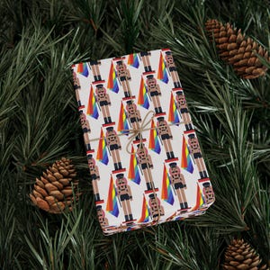 May include: A rectangular gift box wrapped in white paper featuring a repeating pattern of nutcracker figures in black leather-style outfits, each holding a vertical rainbow flag. The package is tied with thin brown twine centered on the top.