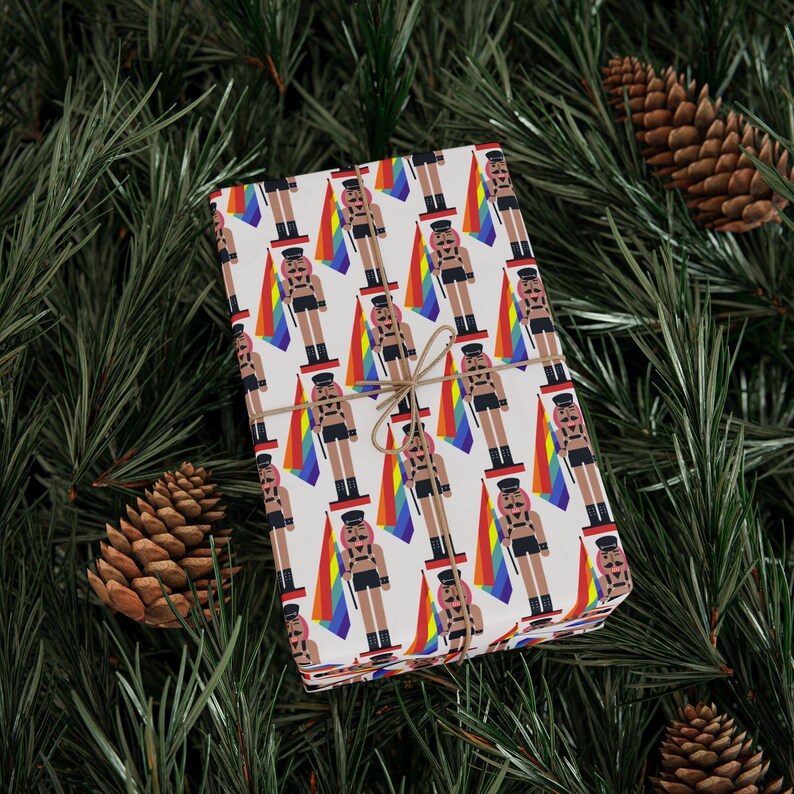 May include: A rectangular gift box wrapped in white paper featuring a repeating pattern of nutcracker figures in black leather-style outfits, each holding a vertical rainbow flag. The package is tied with thin brown twine centered on the top.