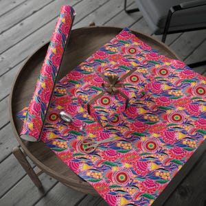 Puede incluir: Un rollo de papel de regalo rosa con un estampado floral colorido, junto a una caja de regalo envuelta y unas tijeras sobre una mesa de madera. El dise&ntilde;o incluye elementos rosas, amarillos, azules y verdes.