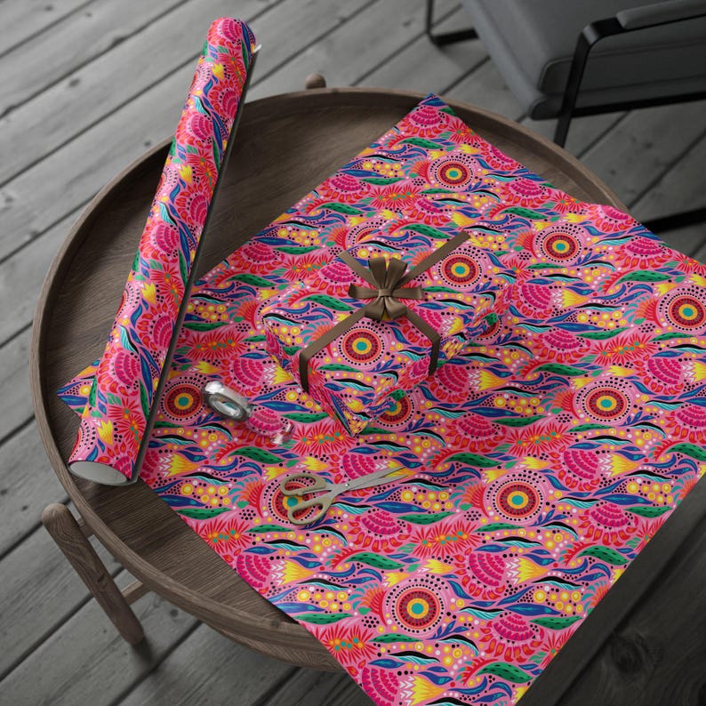 Puede incluir: Un rollo de papel de regalo rosa con un estampado floral colorido, junto a una caja de regalo envuelta y unas tijeras sobre una mesa de madera. El dise&ntilde;o incluye elementos rosas, amarillos, azules y verdes.