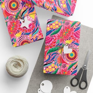 Puede incluir: Tres regalos envueltos con papel estampado floral rosa, atados con hilo y etiquetas blancas. Tambi&eacute;n hay una madeja de hilo, tijeras y etiquetas en blanco. Los regalos est&aacute;n sobre una tela gris y una superficie blanca.