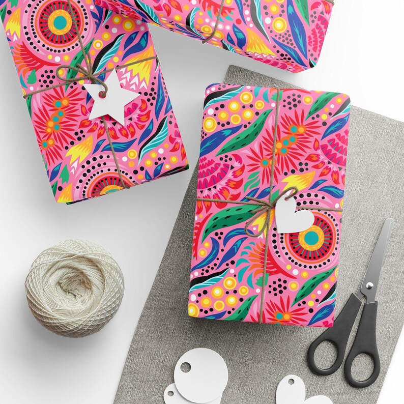 Puede incluir: Tres regalos envueltos con papel estampado floral rosa, atados con hilo y etiquetas blancas. Tambi&eacute;n hay una madeja de hilo, tijeras y etiquetas en blanco. Los regalos est&aacute;n sobre una tela gris y una superficie blanca.
