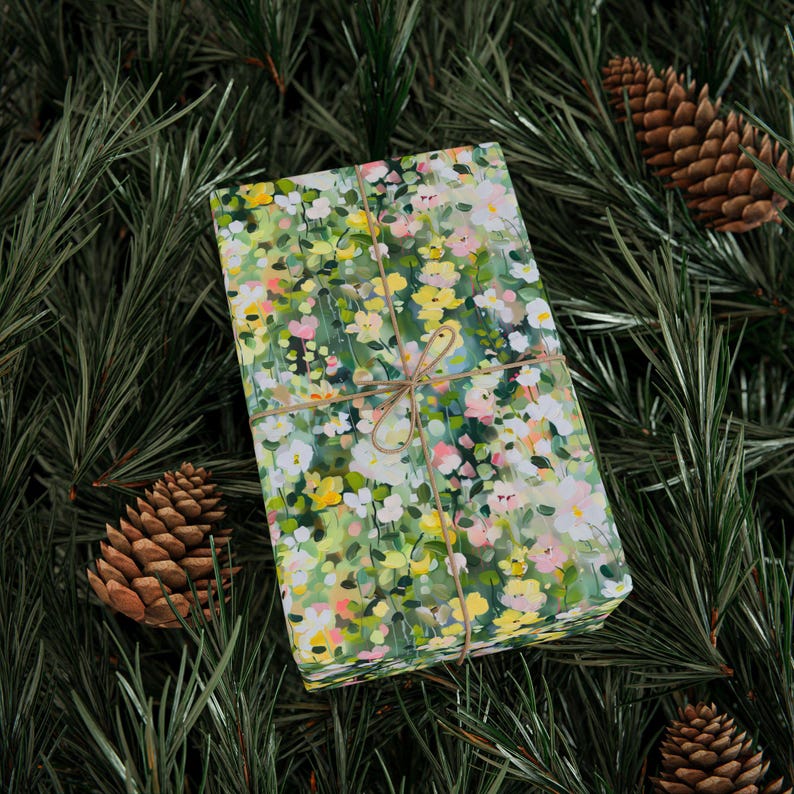 May include: A rectangular gift wrapped in floral patterned paper with yellow, pink, and white flowers on a green background. The gift is tied with twine and rests on a bed of pine needles and pine cones.
