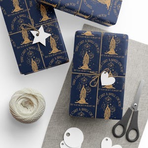 May include: Dark blue wrapping paper featuring a repeating gold pattern of a seated figure with long hair, celestial symbols, and the text "I CAST A SPELL ON YOU BUT YOU WON'T FORGET ME". The paper is tied with twine and white star or heart tags.