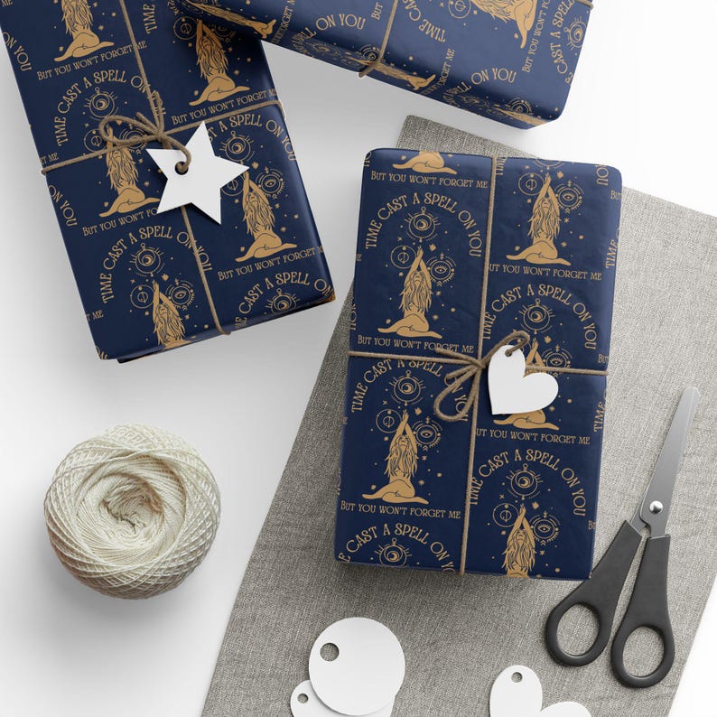 May include: Dark blue wrapping paper featuring a repeating gold pattern of a seated figure with long hair, celestial symbols, and the text "I CAST A SPELL ON YOU BUT YOU WON'T FORGET ME". The paper is tied with twine and white star or heart tags.