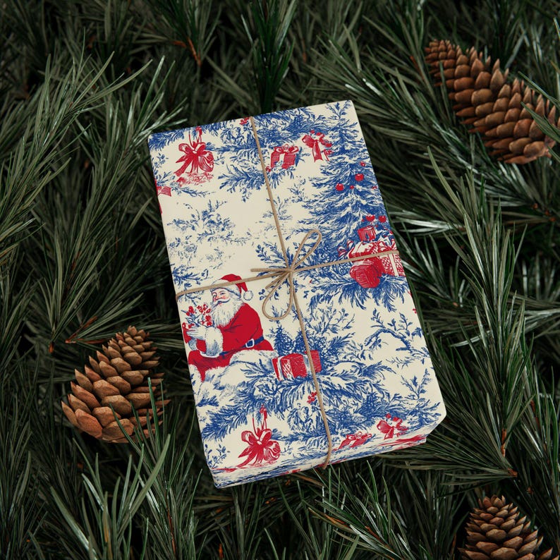May include: A rectangular gift box wrapped in cream-colored paper featuring a blue toile-style pattern of Santa Claus, pine trees, and presents with red accents. The package is tied with thin brown twine centered across the top surface.