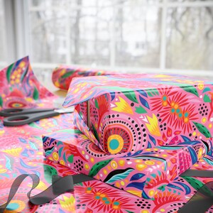 Puede incluir: Cajas de regalo rosas envueltas en papel floral con tijeras y una cinta negra. El papel de regalo presenta un dise&ntilde;o vibrante con elementos florales rojos, amarillos, azules y verdes sobre un fondo rosa. La escena est&aacute; ambientada en una mesa cerca de una ventana.