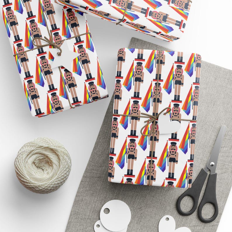 May include: Wrapping paper featuring a repeating pattern of nutcracker figures dressed in black leather-style harnesses and caps, each holding a rainbow flag. The design is printed on a white background and tied with brown twine and star tags.