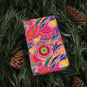 Puede incluir: Un regalo envuelto en papel rosa brillante con un dise&ntilde;o floral, atado con hilo, descansa sobre un lecho de agujas de pino verdes. El dise&ntilde;o colorido incluye flores y hojas amarillas, azules y rojas. Se ven dos pi&ntilde;as.