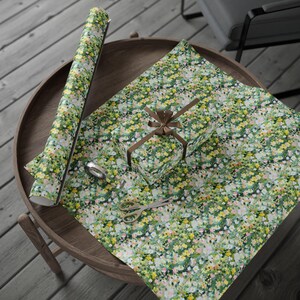 May include: A gift wrapped in floral patterned paper with a brown ribbon, a roll of matching wrapping paper, scissors, and tape on a wooden table. The wrapping paper features a pattern of small yellow, pink, and white flowers on a green background.