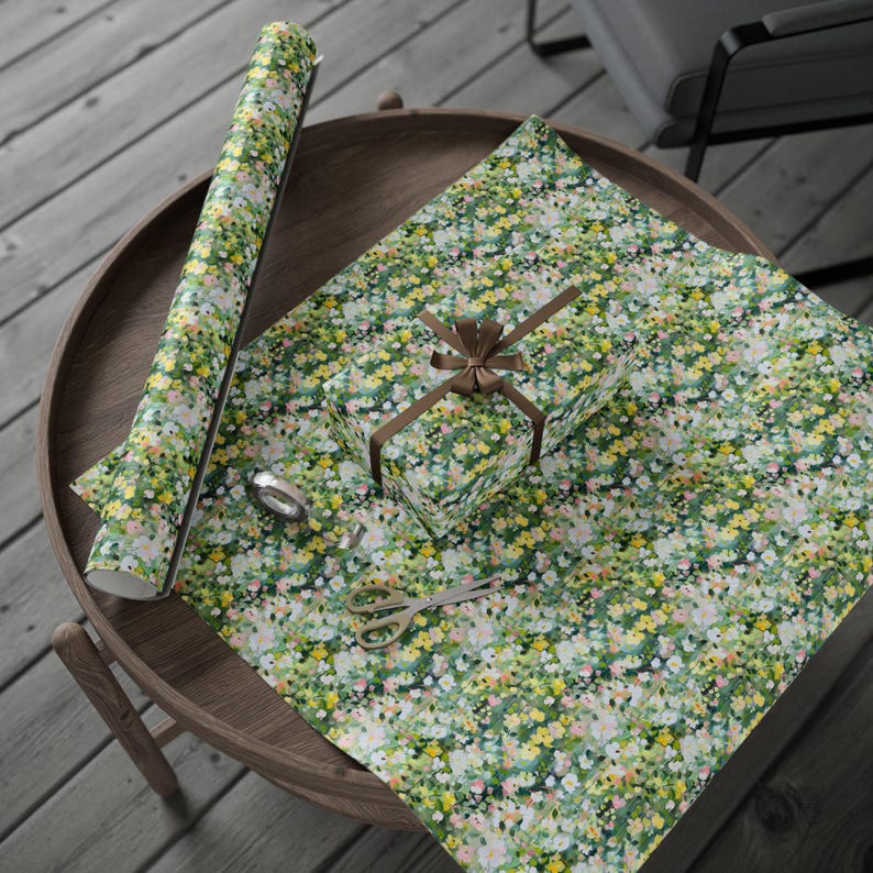 May include: A gift wrapped in floral patterned paper with a brown ribbon, a roll of matching wrapping paper, scissors, and tape on a wooden table. The wrapping paper features a pattern of small yellow, pink, and white flowers on a green background.