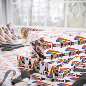 May include: Wrapping paper with a repeating pattern of nutcracker figures wearing black leather-style harnesses and caps, each holding a rainbow-colored flag. The background is white, and the design features vibrant rainbow triangular accents.