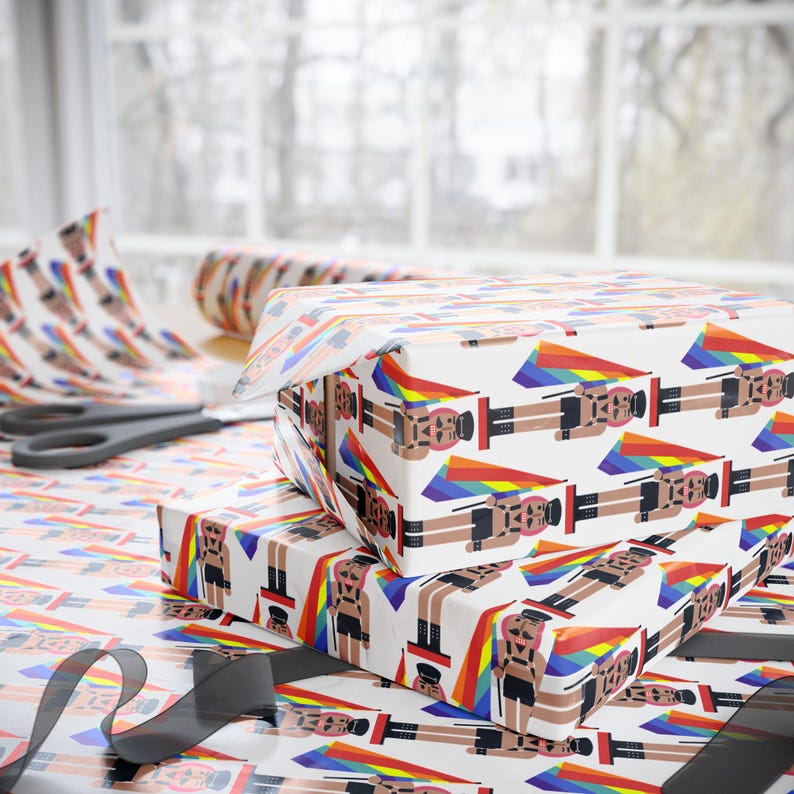 May include: Wrapping paper with a repeating pattern of nutcracker figures wearing black leather-style harnesses and caps, each holding a rainbow-colored flag. The background is white, and the design features vibrant rainbow triangular accents.