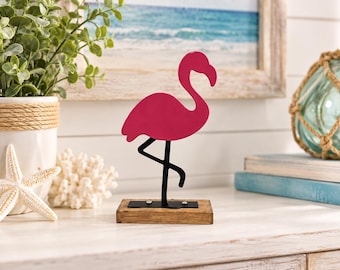 May include: A pink metal flamingo figurine standing on one leg, attached to a rectangular wooden base with two visible screws. The flamingo has a smooth, solid pink body and a black metal leg structure, creating a simple, upright silhouette.