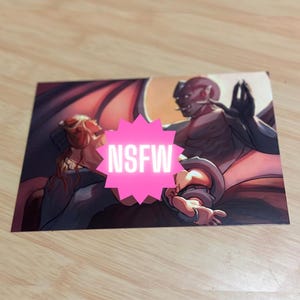 May include: A print featuring a fantasy illustration of two figures, one with red skin and horns, the other with blonde hair. The image has a pink starburst with the text "NSFW" across the center.