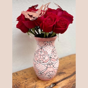 May include: A pink ceramic vase with a floral pattern and black accents. The vase is filled with red roses and decorated with pink butterfly cutouts.