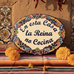 Peut inclure: Panneau ovale en céramique avec le texte "En esta Casa la Reina no Cocina" en écriture bleue, entouré d'une bordure florale colorée. Le panneau repose sur un textile rayé avec des fleurs de souci. Des piments rouges séchés pendent en arrière-plan.