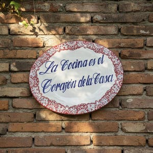 May include: An oval ceramic sign with the text "La Cocina es el Corazón de la Casa" in blue script. The sign has a white background with a red and white decorative border. The sign is mounted on a brick wall.