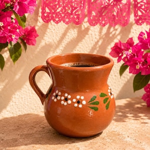 Op de afbeelding: Een bruine, handgemaakte keramische mok gevuld met donkere vloeistof, mogelijk koffie of warme chocolademelk. De mok is versierd met witte en zwarte bloemmotieven en groene bladeren. Heldere roze bloemen en decoratief papier hangen op de achtergrond.
