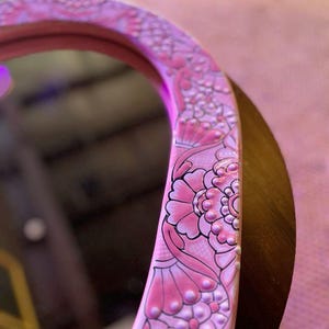 May include: A pink oval mirror with a decorative floral design around the edge. The design features pink and white flowers with black outlines and white dots.