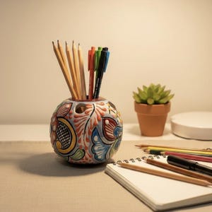 Puede incluir: Un portalápices redondo de cerámica pintado a mano con un diseño floral colorido. El portalápices contiene lápices y bolígrafos. También se ve una pequeña suculenta en maceta y un bloc de notas con lápices.