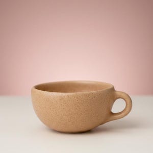 May include: A speckled, tan-colored ceramic teacup with a rounded body and a small, circular handle. The cup sits on a white surface against a soft pink background. The cup's design is simple and modern.