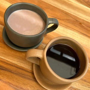 May include: Two ceramic mugs with matching saucers sit on a wooden surface. One mug is filled with a light brown liquid, and the other is filled with a dark brown liquid. Both mugs have a handle and are a similar shape.