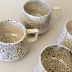 May include: A collection of speckled ceramic mugs. The mugs are off-white with black speckles, featuring a rounded body and a small, rectangular handle. The interior of the mugs is a lighter shade of off-white.