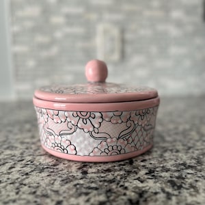 May include: A pink ceramic container with a lid. The container has a floral design in black and white.