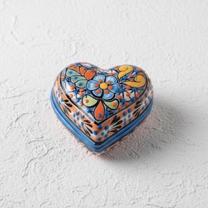 May include: Heart-shaped ceramic trinket box with a blue base and lid. The lid is decorated with a floral pattern in orange, blue, yellow, and green. The box is likely used for storing small items like jewelry or keepsakes.