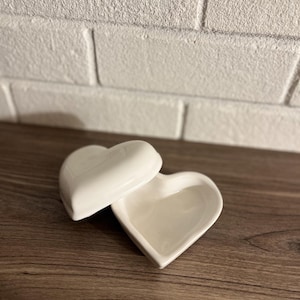 May include: A white ceramic heart-shaped trinket box with a lid, open to reveal the interior. The box sits on a wooden surface, with a white brick wall in the background. The box is designed to hold small items.