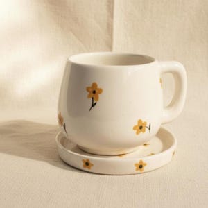 May include: Cream-coloured ceramic mug and saucer set. The mug has a rounded shape with a handle and is decorated with small yellow flowers. The saucer also features the same floral pattern. Ideal for serving hot drinks.
