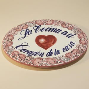 May include: Oval ceramic decorative plate with a white background, featuring a red heart and the phrase "La cocina es el corazón de la casa" in blue script. The border has a pink floral design.