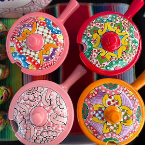 Könnte beinhalten: Vier farbenfrohe Keramik-Servierplatten mit Deckeln und Griffen. Jede Platte zeigt ein anderes Blumenmuster in den Farben Rosa, Rot, Gelb, Grün und Blau.