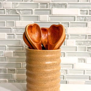 Könnte beinhalten: Ein Set aus hölzernen herzförmigen Löffeln in einem beigefarbenen Keramik-Utensilienhalter. Die Löffel haben eine warme braune Farbe, und der Halter hat ein strukturiertes, geriffeltes Design. Der Hintergrund zeigt einen weißen und grauen Fliesenspiegel.