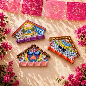 May include: Three colourful ceramic key holders shaped like houses, each with a row of hooks. The key holders feature intricate floral and geometric patterns in vibrant colours. Pink paper banners and bougainvillea flowers decorate the background.