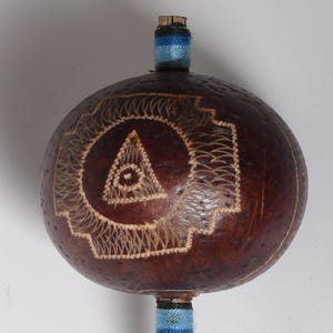 May include: A round, brown gourd with a carved design featuring a triangle with an eye inside a circle. The gourd has a wooden stopper and blue and black striped accents. The gourd is a decorative item.