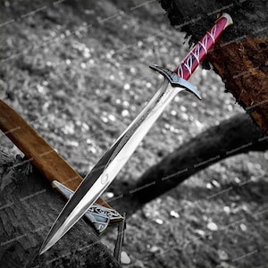 May include: A silver sword with a polished blade and a decorative hilt. The handle is red with white accents. The sword is resting on a wooden surface, with a brown sheath visible.