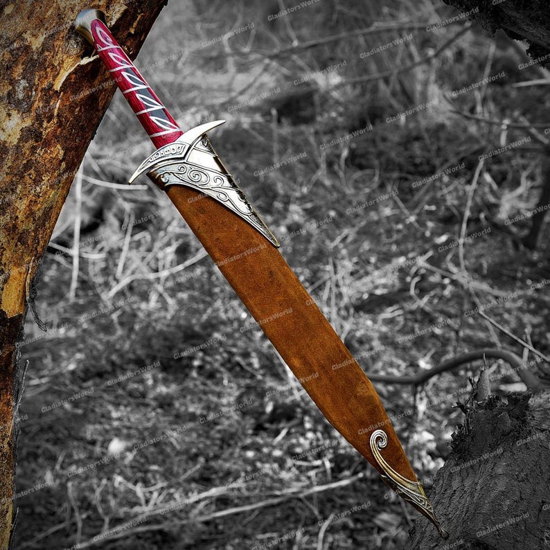 May include: A decorative sword with a brown sheath and a red and silver handle. The sword has a silver guard with an ornate design. The blade is not visible. The sword is leaning against a tree in a black and white outdoor setting.