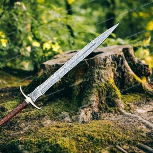 May include: A Damascus steel sword with a patterned blade, a brown wooden handle, and a gold-colored hilt. The sword is displayed outdoors, resting on a moss-covered tree stump, with a blurred green background.