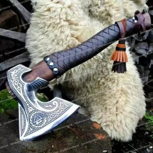 May include: A silver and wood axe with a decorative blade featuring intricate patterns. The handle is wrapped in dark leather with a braided design and a brown leather tassel. The axe is displayed on a wooden surface.