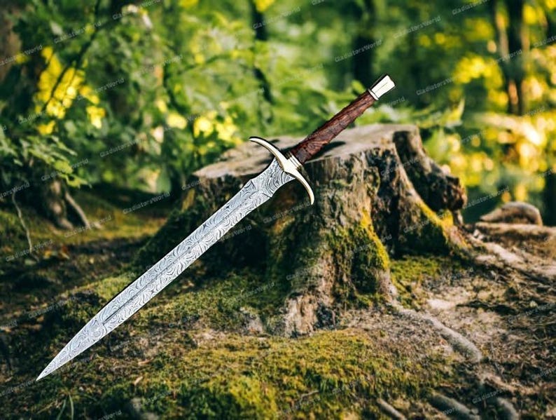 May include: A detailed, silver-colored sword with a patterned blade and a brown handle. The sword is resting on a moss-covered tree stump in a forest setting, with green foliage in the background.