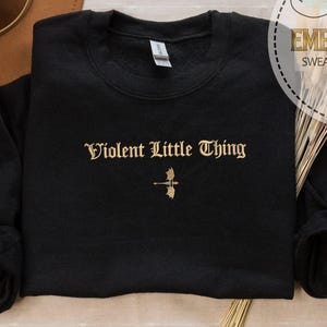 May include: Black crewneck sweatshirt with the embroidered text "Violent Little Thing" in gold lettering. A small gold graphic is below the text. A brown hat is in the background. The sweatshirt is part of a personalized gift collection.