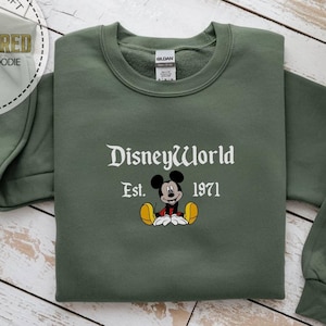 May include: An olive green sweatshirt featuring the embroidered text "DisneyWorld" and "Est. 1971" in white. A cartoon Mickey Mouse image is embroidered below the text. The sweatshirt is folded on a light wood surface.