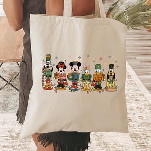 May include: A cream-colored tote bag featuring a colorful illustration of cartoon characters in a row. The characters are dressed in festive outfits. The bag has two long handles and is being held by a person.