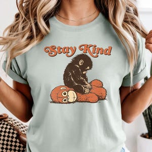 May include: A light green t-shirt featuring a graphic of a small, dark-colored monkey sitting on a red-orange teddy bear. The text "Stay Kind" is arched above the monkey and bear illustration in orange.