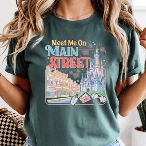 May include: Dark green t-shirt with the text "Meet Me On Main Street" in blue and red. The graphic includes a castle, buildings, and fireworks. The shirt is a short-sleeved crew neck.