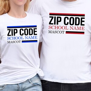May include: Two white t-shirts with the text "ZIP CODE SCHOOL NAME MASCOT" printed in black, blue, and red. The shirts are worn by two people. The background is a red wooden wall.
